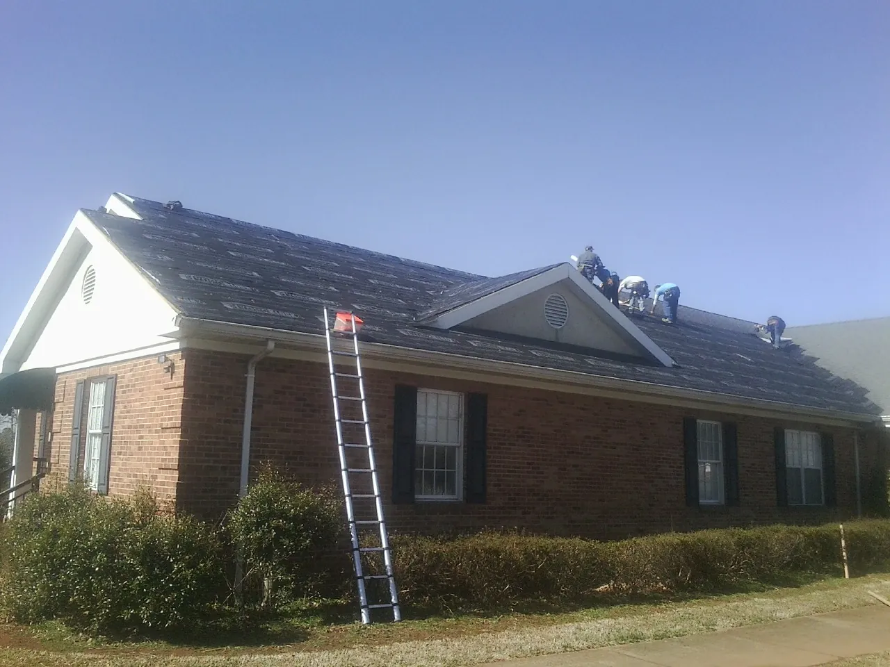 Crew tearing shingles off a brick ranch roof with a ladder set — Emergency Roof Repair work by our crew in Oldhams