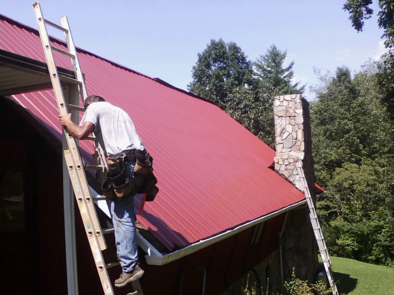 Roofer on a ladder beside a red metal roof and stone chimney — Soffit & Fascia Repair by Roofing Brothers in Oldhams